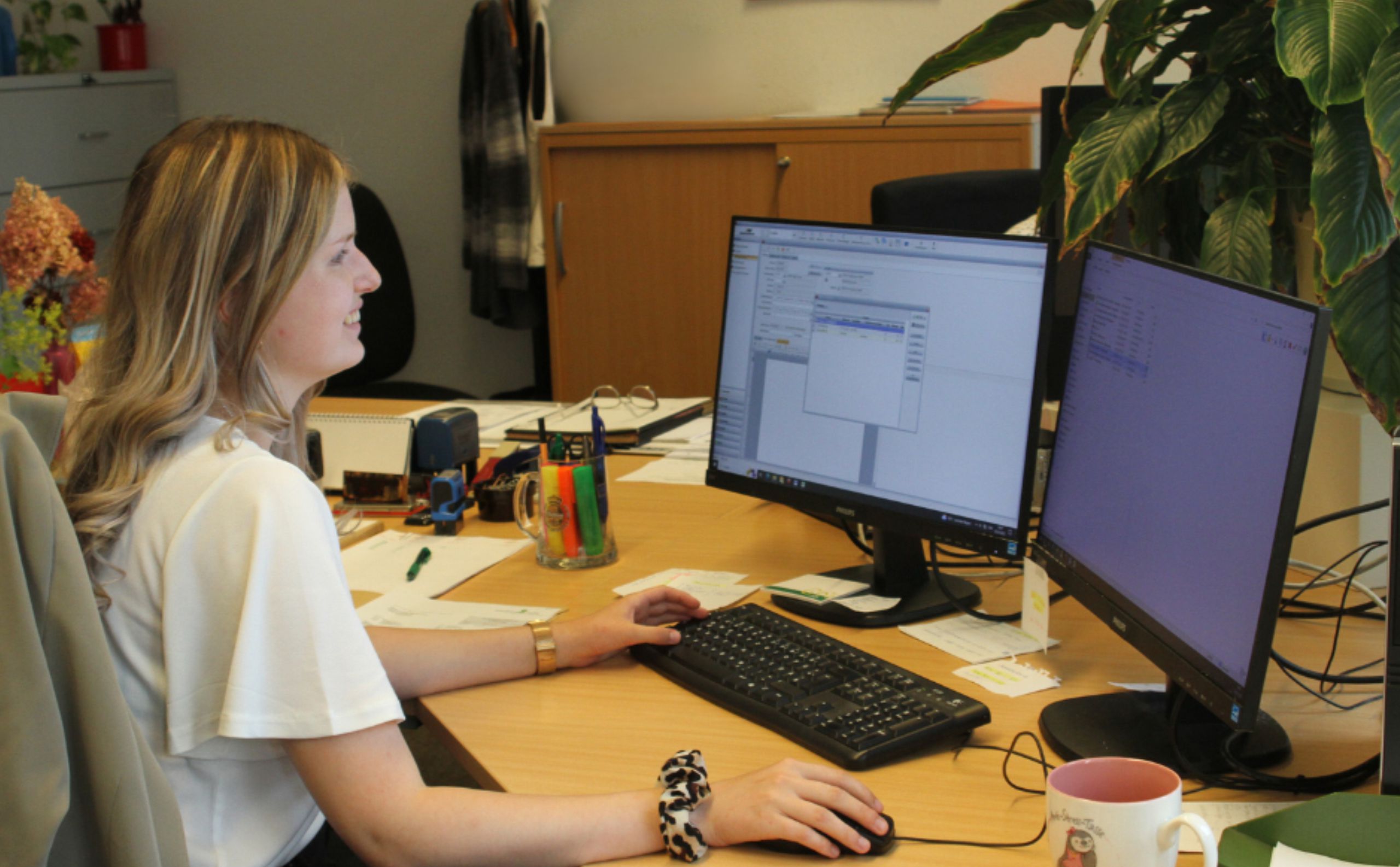 Ausbildung-Industriekaufleute – Feinguss Lobenstein Eine Frau sitzt an einem Schreibtisch und arbeitet konzentriert an zwei Computerbildschirmen. Die Szene zeigt Büroarbeit in einer organisierten und angenehmen Arbeitsumgebung.
